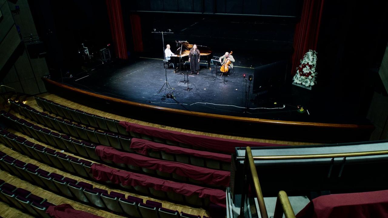 Blick vom ersten Rang eines leeren Opernhauses zu drei Musikern auf der Bühne.