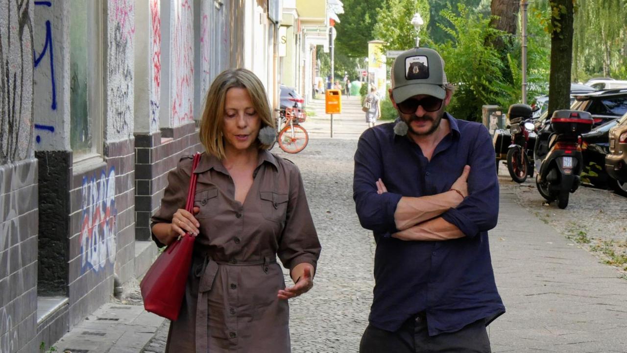 Gisela Steinhauer läuft links von Milan Peschel auf einem Bürgersteig, beide schauen nach unten