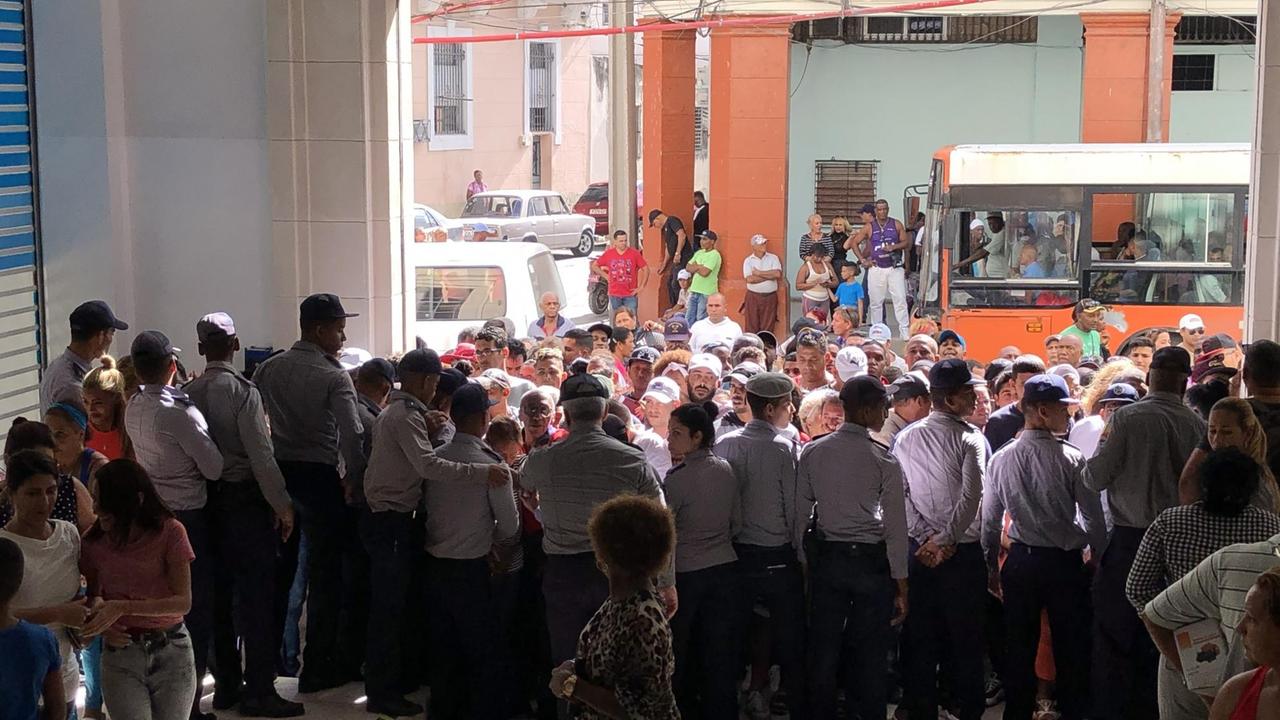 In einen breiten Gebäudeeingang drängeln viele Menschen auf einmal. Im Hintergrund hält ein Bus. Unmittelbar vor den drängelnden Menschen stehen dicht an dicht Polizeibeamte in einer Reihe und hindern sie am Eintreten. In einen breiten Gebäudeeingang drängeln viele Menschen auf einmal. Im Hintergrund hält ein Bus. Unmittelbar vor den drängelnden Menschen stehen dicht an dicht Polizeibeamte in einer Reihe und hindern sie am Eintreten.