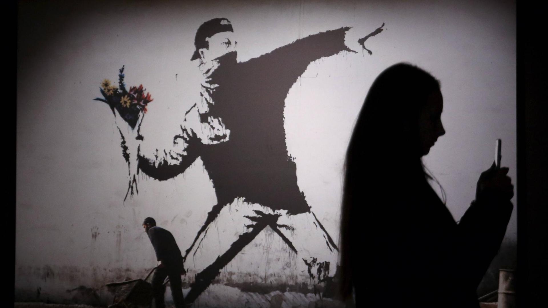 Das Bild zeigt die Schatten von Besuchern einer Banksy-Ausstellung in St. Petersburg, Russland. Zu sehen ist das Bild "The Flower Thrower" - der Blumenwerfer, das stilisierte Abbild eines Protestierenden, der mit seinem Arm weit ausholt, um einen Strauß Blumen zu werfen.