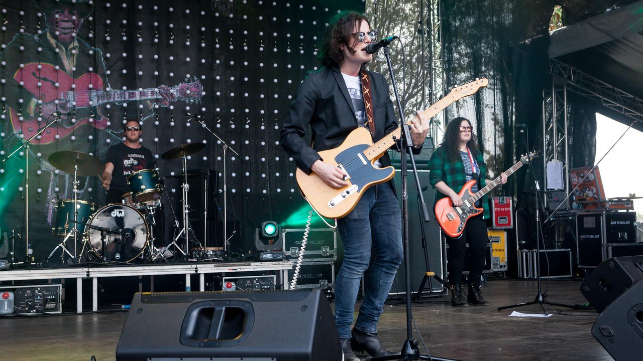 Eine Frau (Bass) und zwei Männer (Gitarre/Gesang und Schlagzeug) stehen auf einer beleuchteten Konzertbühne. Eine Frau (Bass) und zwei Männer (Gitarre/Gesang und Schlagzeug) stehen auf einer beleuchteten Konzertbühne.