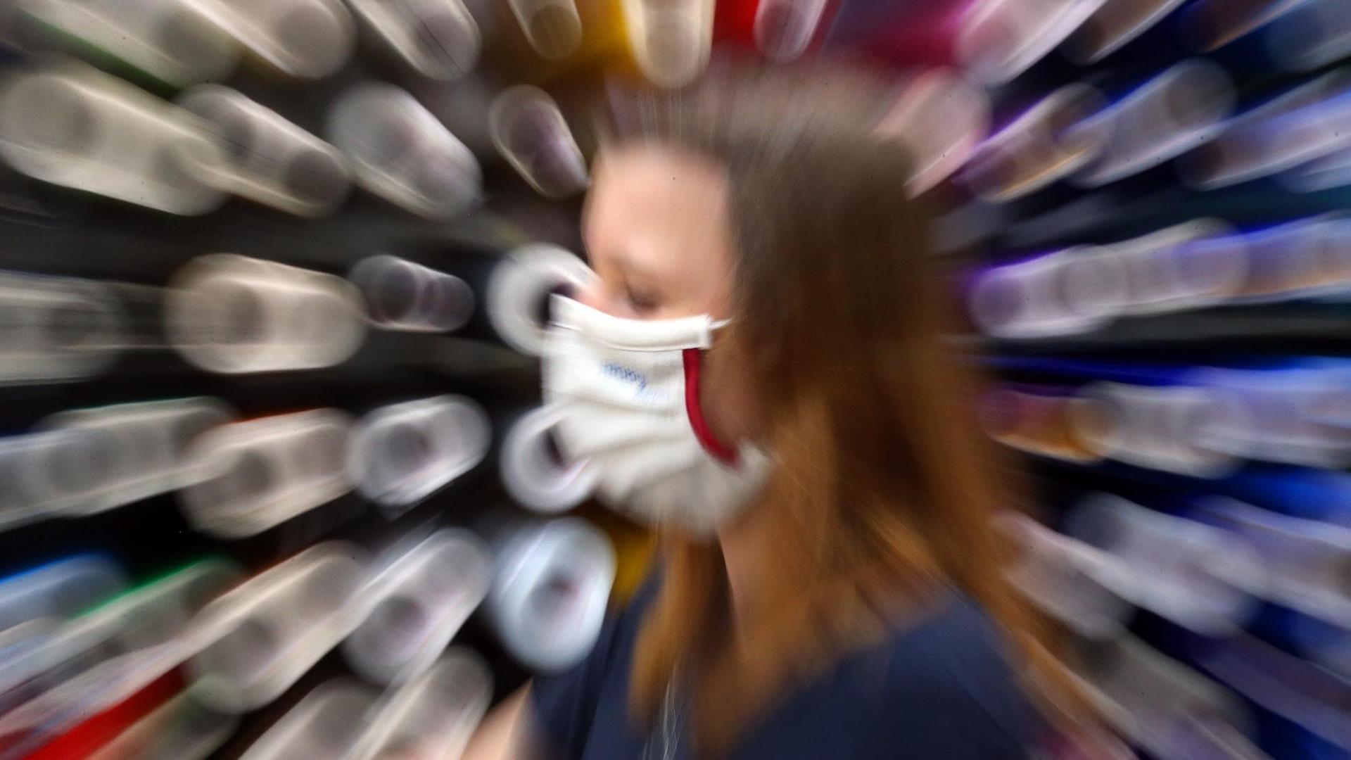 Mit der Mund- und Nasen-Bedeckung als ständigem Begleiter geht eine junge Frau an aufgestapelten Papierrollen entlang. Der Hintergrund ist unscharf.