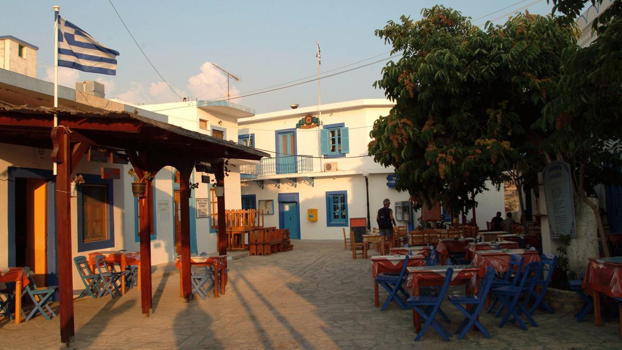 Dorfplatz von Ormos Sokoro mit griechischer Fahne und einem leeren Straßencafe. Dorfplatz von Ormos Sokoro mit griechischer Fahne und einem leeren Straßencafe.