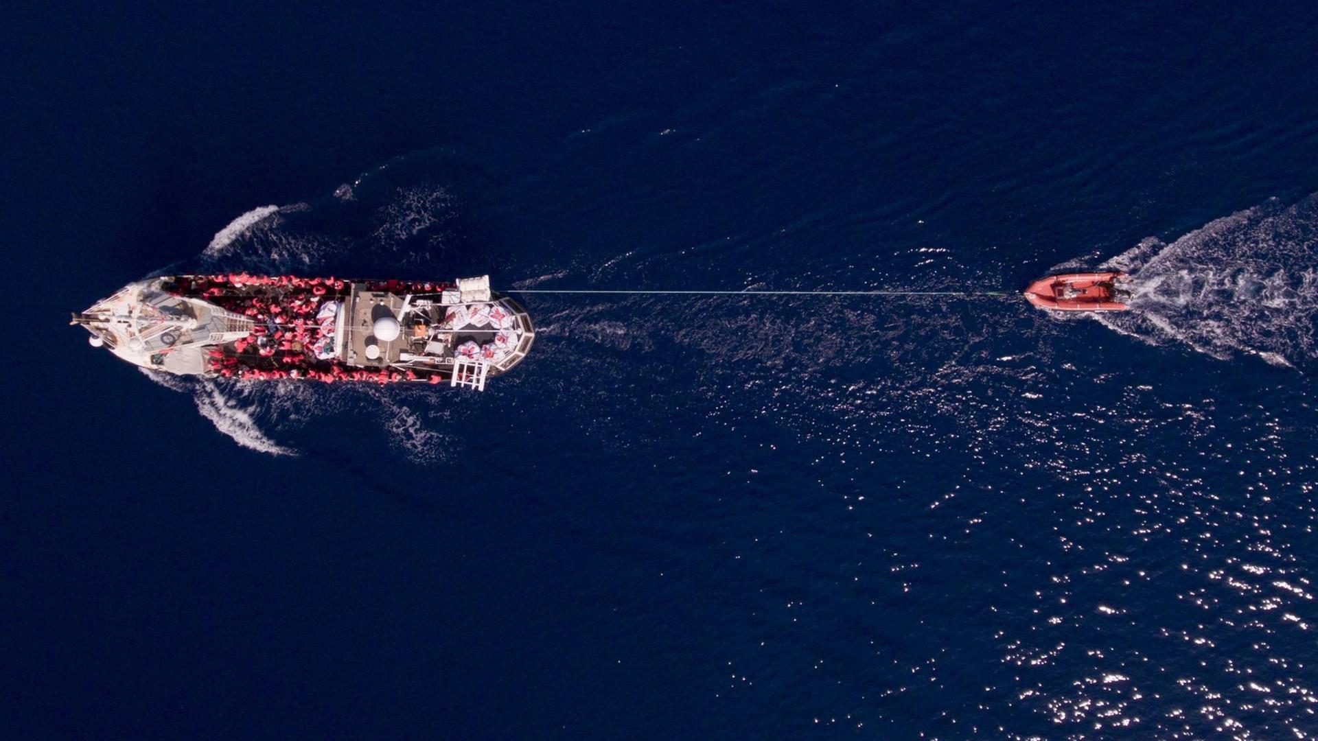 Luftaufnahme auf das Rettungsschiff „Eleonore“ welches ein Schlauchboot hinter sich her zieht. Auf dem Boot sind Menschen in Rettungswesten zu sehen. Luftaufnahme auf das Rettungsschiff „Eleonore“ welches ein Schlauchboot hinter sich her zieht. Auf dem Boot sind Menschen in Rettungswesten zu sehen.