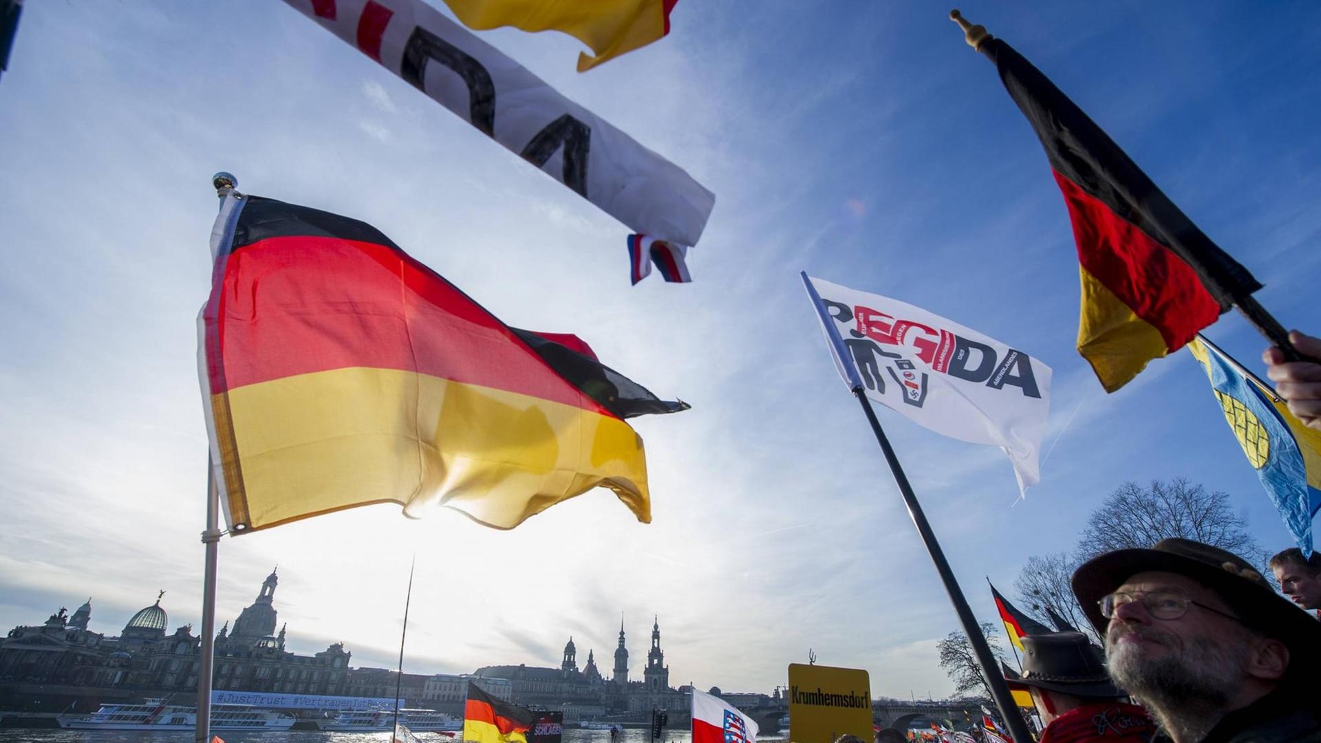 Deutschland-Fahnen und Pegida-Fahnen wehen im Wind vor blauem Himmel, am unteren Bildrand ist die Dresdener Altstadtkulisse zu sehen und Menschen, die demonstrieren. Deutschland-Fahnen und Pegida-Fahnen wehen im Wind vor blauem Himmel, am unteren Bildrand ist die Dresdener Altstadtkulisse zu sehen und Menschen, die demonstrieren.