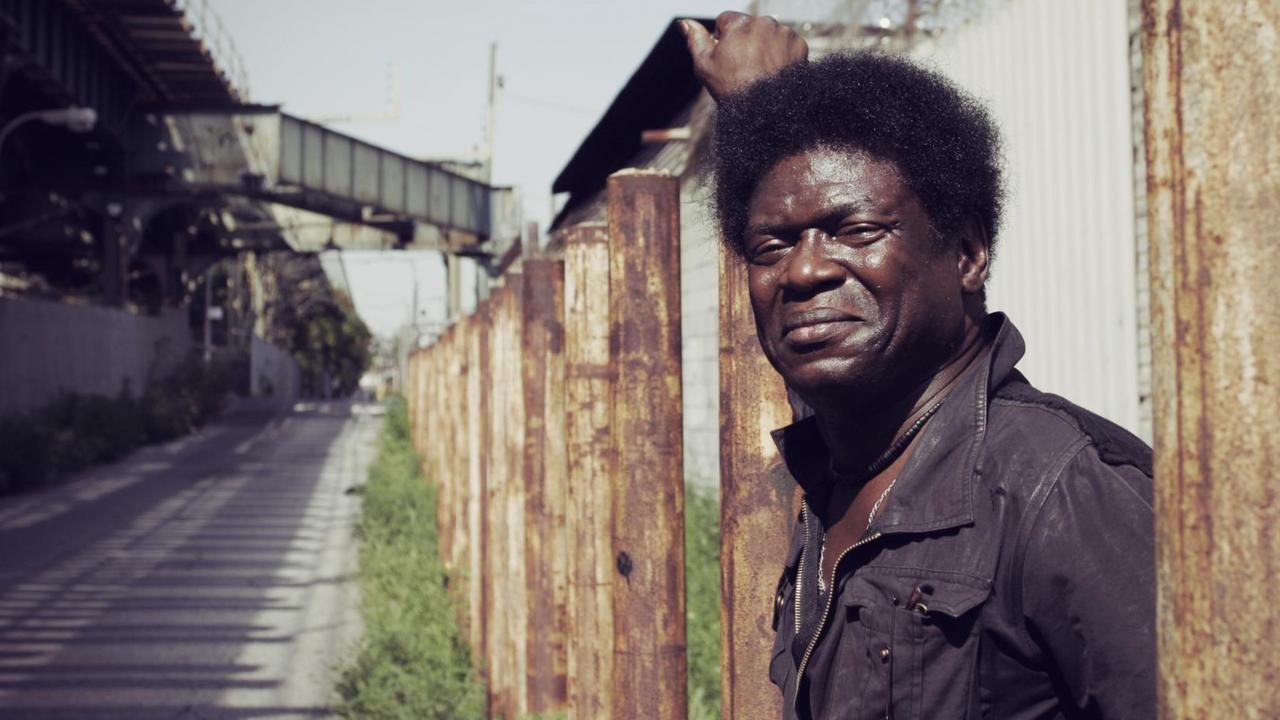 Charles Bradley in einer Tagszene einer Gasse: Er steht an einem Zaun und blickt freundlich in die Kamera. Charles Bradley in einer Tagszene einer Gasse: Er steht an einem Zaun und blickt freundlich in die Kamera.