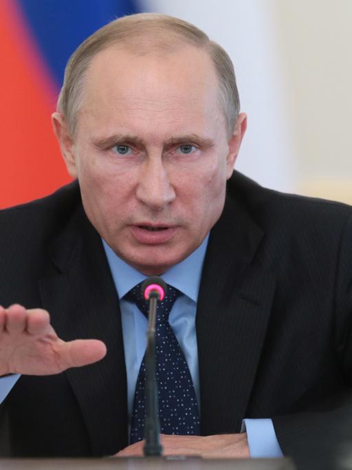Wladimir Putin sitzt an einem Mikrofon, gestikuliert und spricht