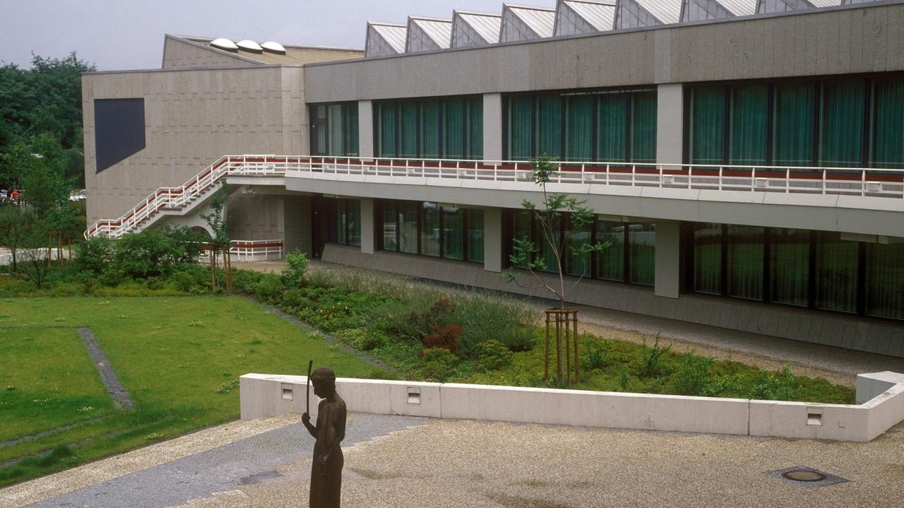 Hohe Fensterfronten eines Gebäudes öffnen sich zu einer Grünfläche mit moderner Figur. Hohe Fensterfronten eines Gebäudes öffnen sich zu einer Grünfläche mit moderner Figur.