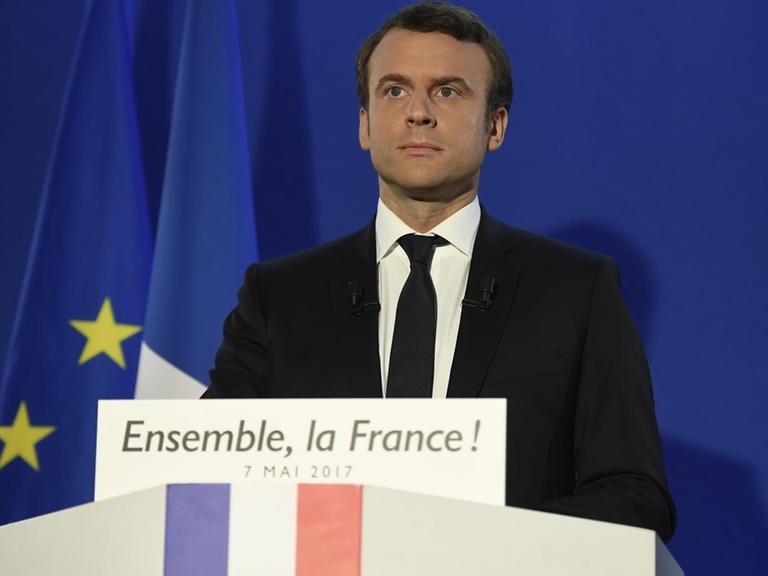 Macron steht mit ernstem Blick vor einer blauen Wand mit Frankreich-Fahne an einem Rednerpult. Auf einem Schild darauf steht "Ensemble, la France!".