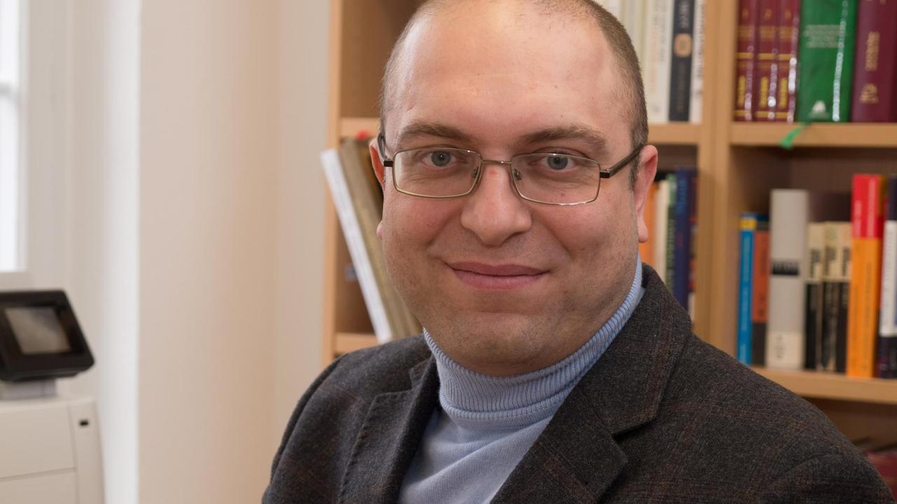 Ein kahler Mann in blauem Rollkragenpullover, dunklem Sakko und mit Brille lächelt freundlich in die Kamera. - im Hintergrund steht ein Bücherregal. Ein kahler Mann in blauem Rollkragenpullover, dunklem Sakko und mit Brille lächelt freundlich in die Kamera. - im Hintergrund steht ein Bücherregal.