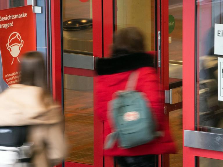 Schüler und Schülerinnen der Städtischen Integrierten Gesamtschule Paffrath gehen durch den Eingang der Schule, an dessen Scheiben Hinweiszettel kleben, die auf die Maskenpflicht und die Abstandsregeln hinweisen.