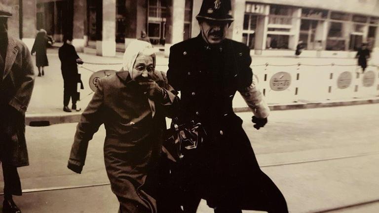 Historische Postkarte: Eine alte Frau hält sich ängstlich an einem Polizisten fest, der versucht, sie über die Straße zu begleiten