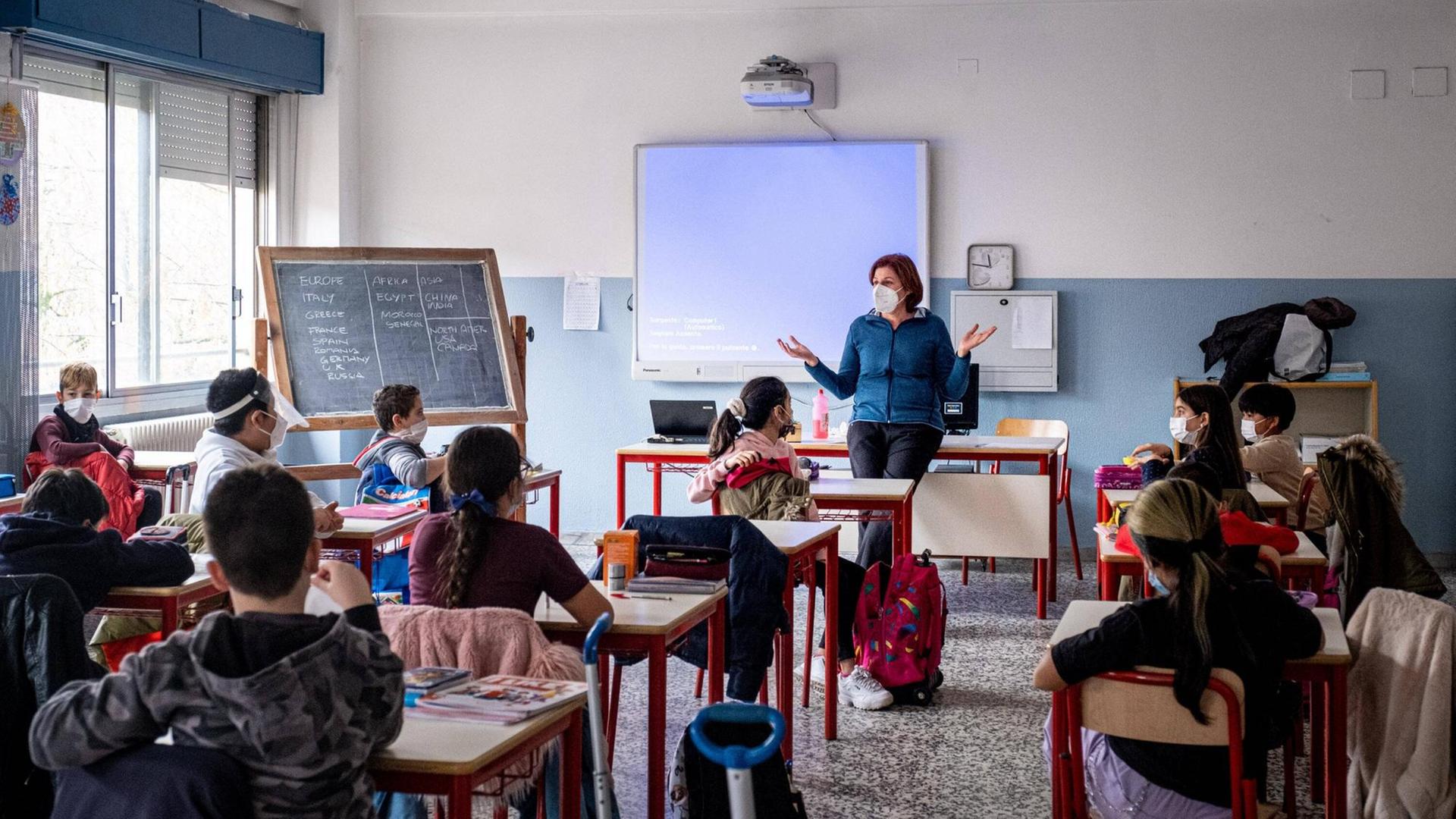 Schülerinnen und Schüler sitzen mit Masken und Abständen in einem Klassenraum. Vorne steht eine Lehrerin, ebenfalls mit Maske. Schülerinnen und Schüler sitzen mit Masken und Abständen in einem Klassenraum. Vorne steht eine Lehrerin, ebenfalls mit Maske.