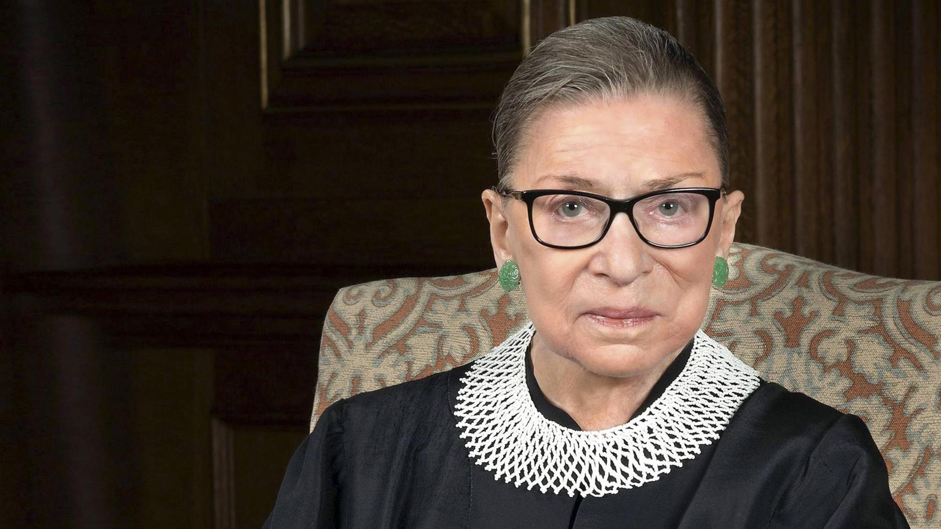 Ruth Bader Ginsburg trägt über ihre schwarze Robe eine weißen geflochtenen Kragen, sie schaut ernst in die Kamera Ruth Bader Ginsburg trägt über ihre schwarze Robe eine weißen geflochtenen Kragen, sie schaut ernst in die Kamera