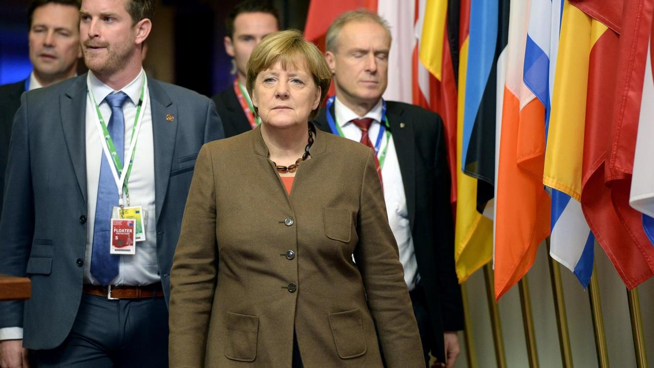 Die Kanzlerin läuft im braunen Jackett an einer Reihe Fahnen der EU-Mitgliedsstaaten vorbei. Hinter ihr laufen vier Security-Leute.