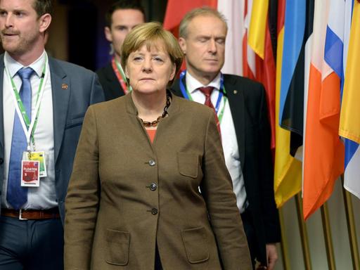 Die Kanzlerin läuft im braunen Jackett an einer Reihe Fahnen der EU-Mitgliedsstaaten vorbei. Hinter ihr laufen vier Security-Leute.