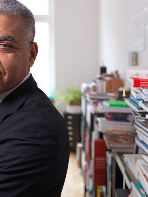 Rayan Abdullah, Professor für Typografie an der Hochschule für Grafik und Buchkunst Leipzig, steht in einem Büro vor einem Regal mit Stapeln von Büchern.