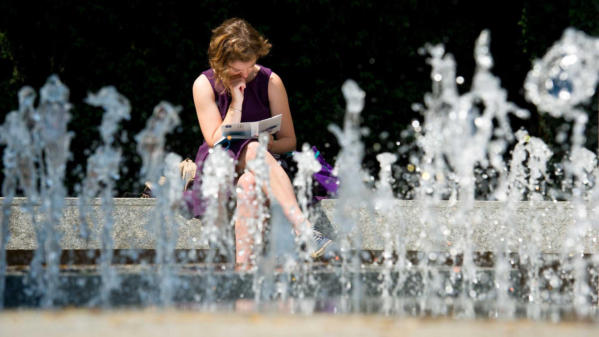 Eine junge Frau sitzt bei Sonnenschein in einem kleinen Park in Frankfurt am Main (Hessen) an einem sprudelnden Brunnen und liest dabei ein Buch.