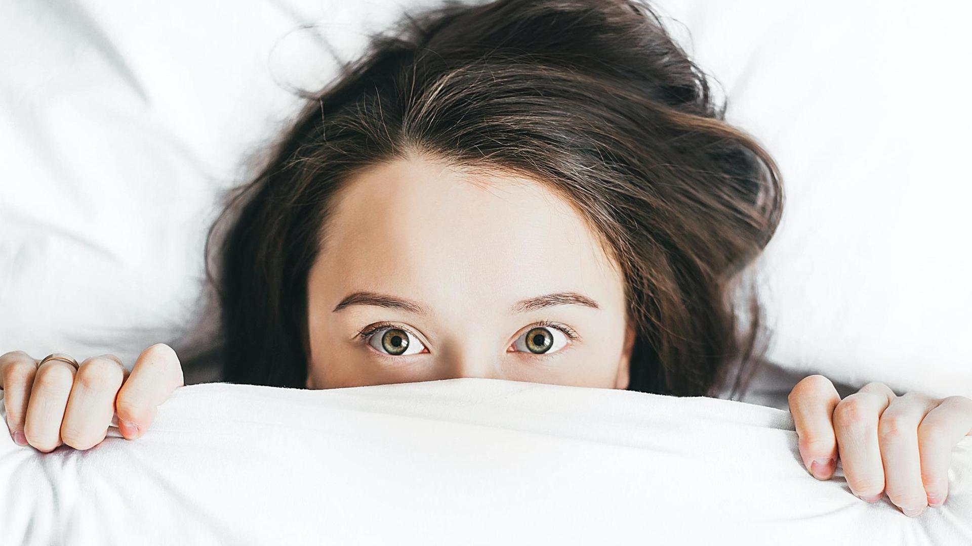 Eine junge Frau mit weit aufgerissenen Augen zieht die Bettdecke halb über ihr gesicht. Eine junge Frau mit weit aufgerissenen Augen zieht die Bettdecke halb über ihr gesicht.