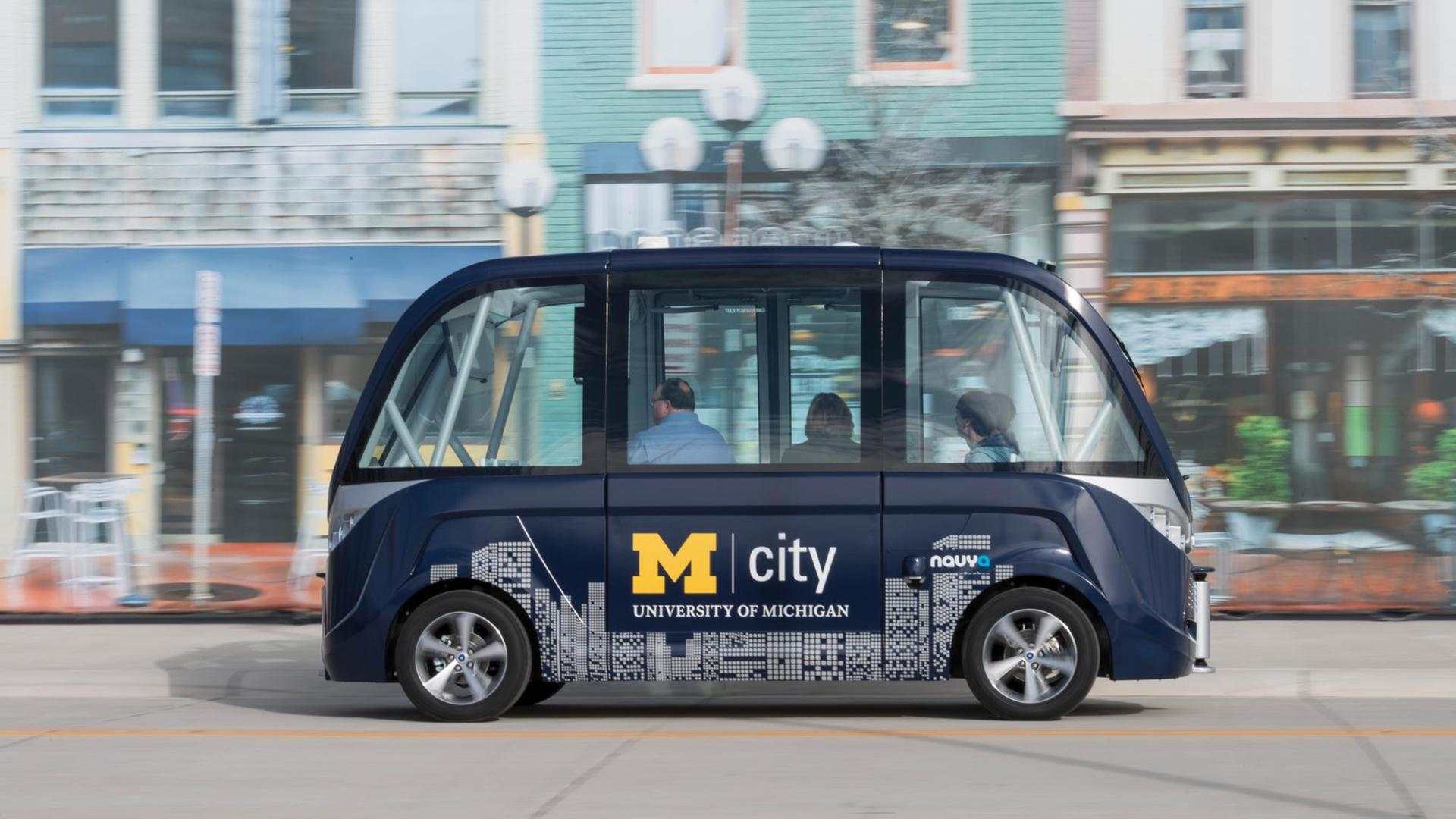 Vor einer Häuserkulisse fährt ein kleiner blauer Bus mit drei Personen jedoch ohne Fahrer vorbei. Der Bus ist mit M City University of Michigan beschriftet.