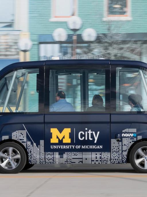 Vor einer Häuserkulisse fährt ein kleiner blauer Bus mit drei Personen jedoch ohne Fahrer vorbei. Der Bus ist mit M City University of Michigan beschriftet.