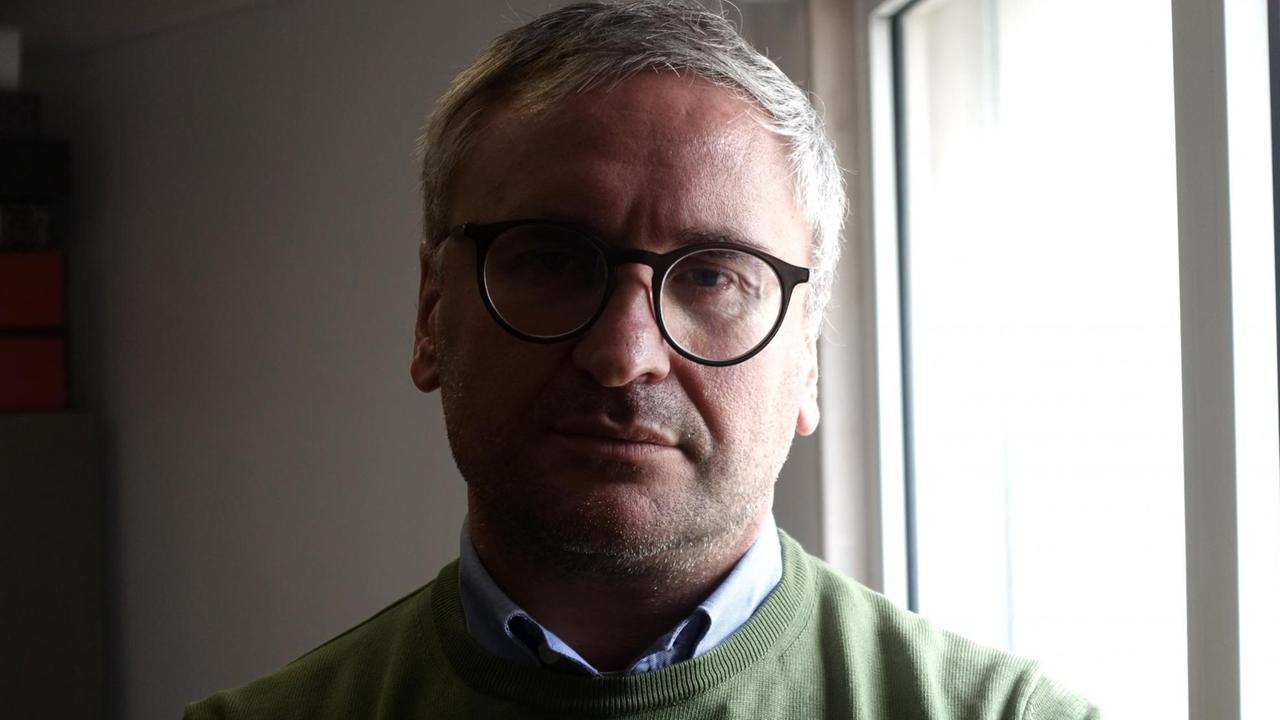 Ein Mann mit kurzen grauen Haaren und Brille sowie olivgrünem Pulli steht in einer Wohnung am Fenster. Ein Mann mit kurzen grauen Haaren und Brille sowie olivgrünem Pulli steht in einer Wohnung am Fenster.