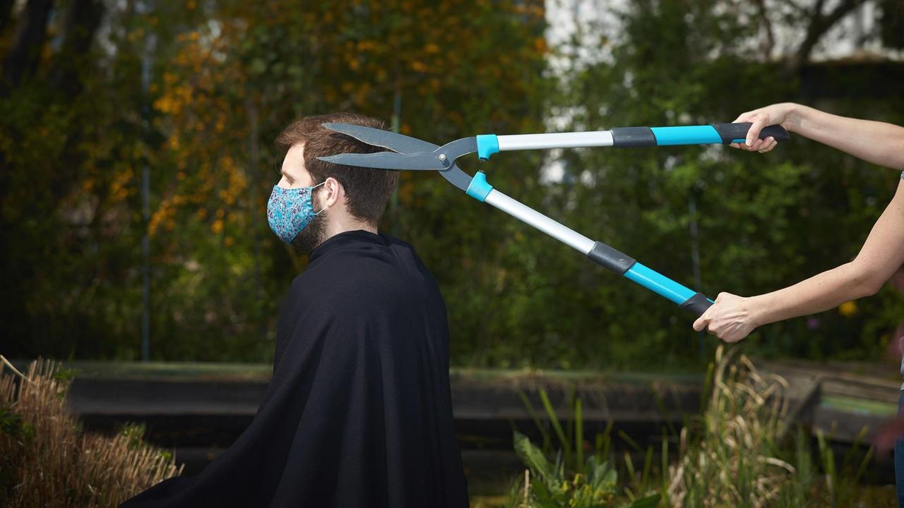 Ein Mann mit Mundschutz bekommt die Haare mit einer Heckenschere geschnitten. Ein Mann mit Mundschutz bekommt die Haare mit einer Heckenschere geschnitten.