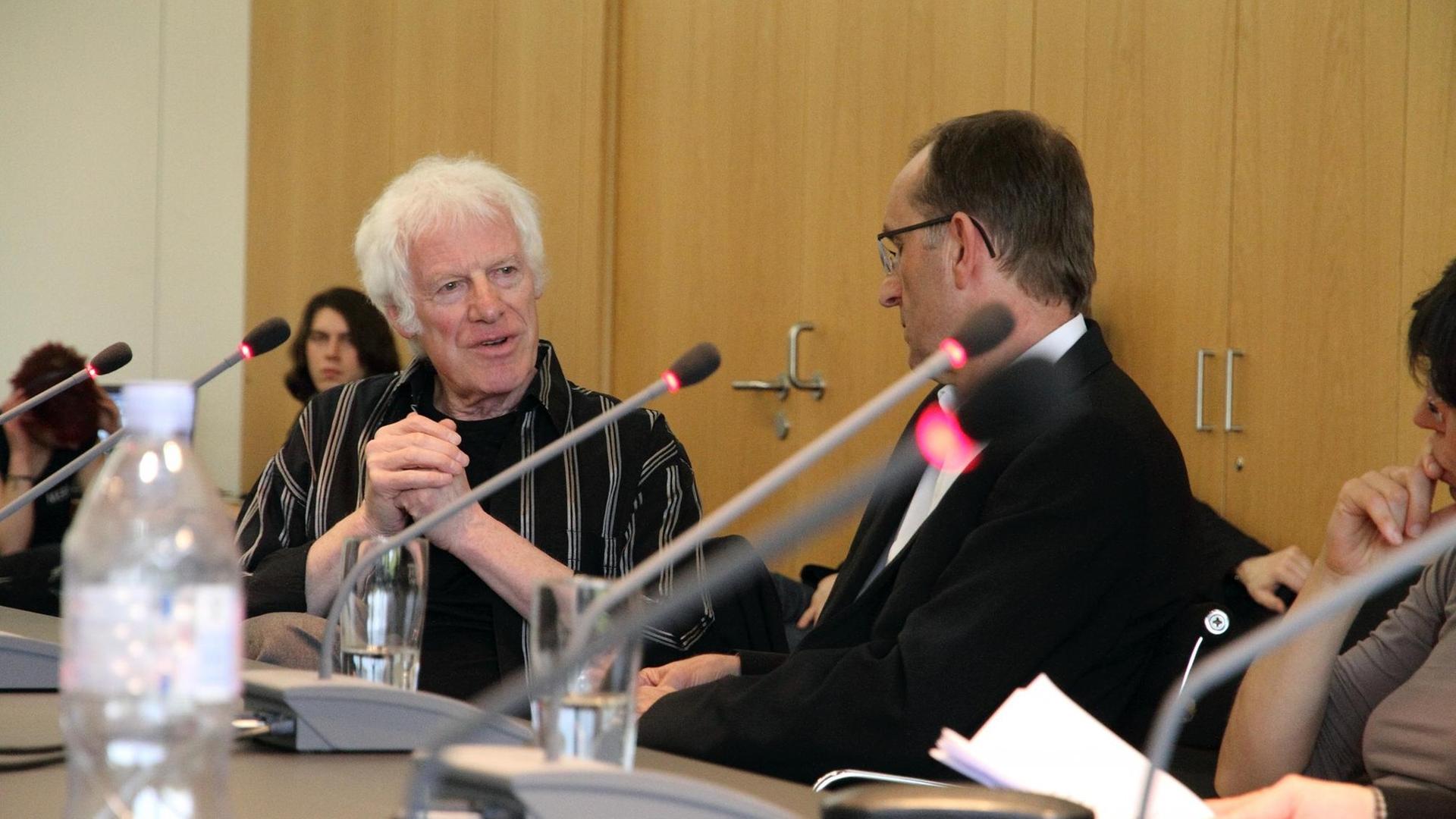 Ein weißhaariger Mann mit einem schwarz-silbern gestreiftem Hemd sitzt an einem Konferenztisch auf dem Mikrofone und Wassergläser stehen und spricht mit einem dunkelhaarigen Mann, der neben ihm sitzt. Ein weißhaariger Mann mit einem schwarz-silbern gestreiftem Hemd sitzt an einem Konferenztisch auf dem Mikrofone und Wassergläser stehen und spricht mit einem dunkelhaarigen Mann, der neben ihm sitzt.