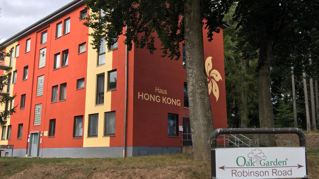 Zwischen Bäumen steht ein rot und gelb angemalertes Mehrfamilienhaus mit der Aufschrift "Haus Hong Kong". Im Vordergrund ein Hinweisschild mit der Aufschrift "Oak Garden, Robinson Road" ("Eichengarten, Robinson-Straße") Zwischen Bäumen steht ein rot und gelb angemalertes Mehrfamilienhaus mit der Aufschrift "Haus Hong Kong". Im Vordergrund ein Hinweisschild mit der Aufschrift "Oak Garden, Robinson Road" ("Eichengarten, Robinson-Straße")