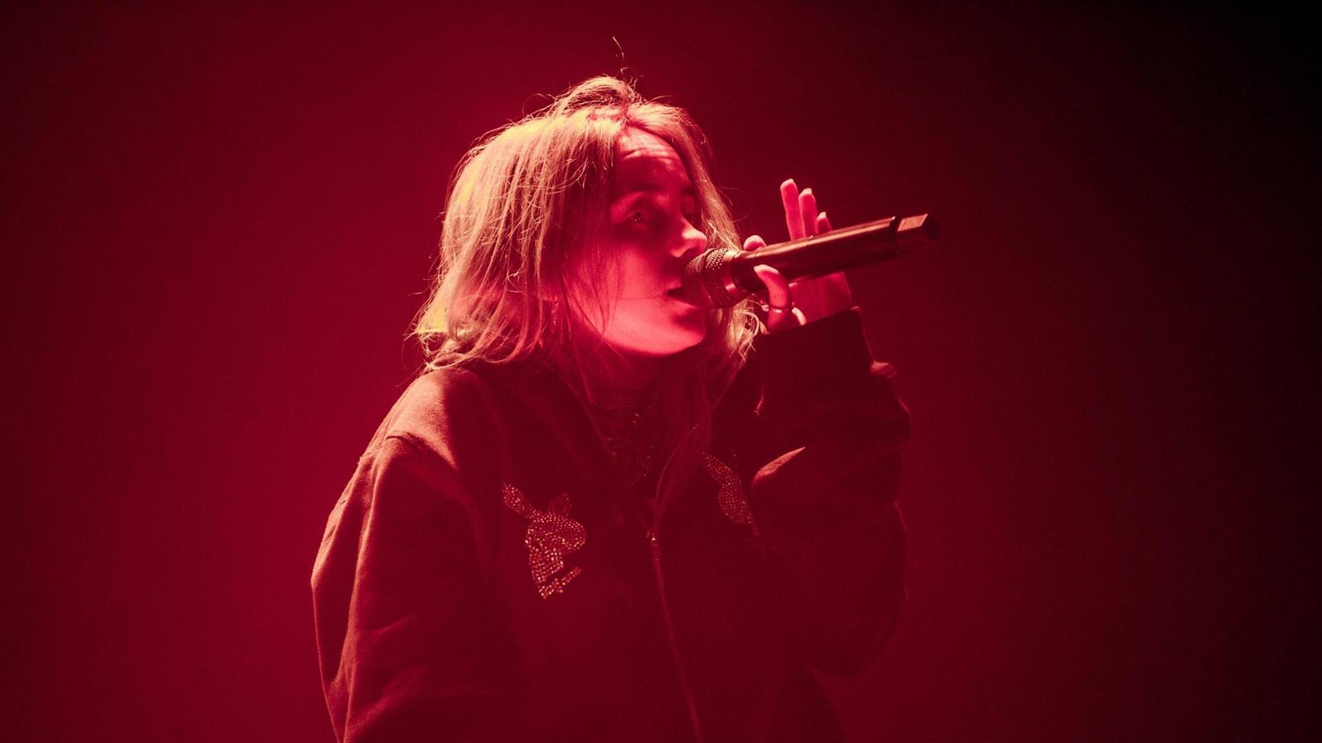 Die junge Sängerin Billie Eilish steht in rotem Licht auf der Bühne und singt mit einem Mikrofon in der linken Hand. Die junge Sängerin Billie Eilish steht in rotem Licht auf der Bühne und singt mit einem Mikrofon in der linken Hand.
