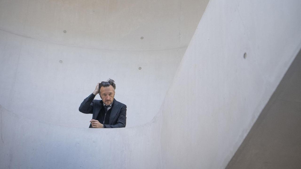 Das Treppenhaus ist aus hellem Beton und bildet mit seinen Linien einen schlichten Hintergrund für Lukas Bärfuss, der mit einem Arm auf dem Geländer aufgestützt ist und sich mit der anderen Hand durch die Haare fährt. Das Treppenhaus ist aus hellem Beton und bildet mit seinen Linien einen schlichten Hintergrund für Lukas Bärfuss, der mit einem Arm auf dem Geländer aufgestützt ist und sich mit der anderen Hand durch die Haare fährt.