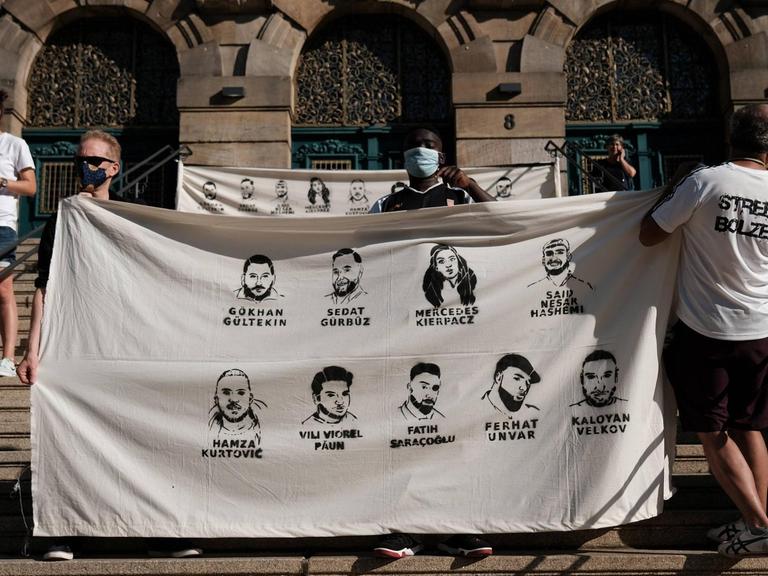 Teilnehmer der Gedenkdemo halten auf Stufen ein Plakat mit den Namen und Gesichtern der Opfer von Hanau