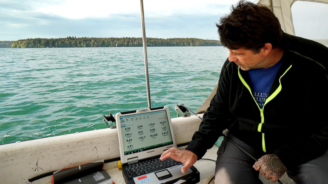 Ein Mann sitzt in einem Boot auf einem See. Vor ihm steht ein Computer. Ein Mann sitzt in einem Boot auf einem See. Vor ihm steht ein Computer.
