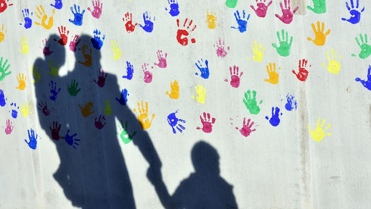 Ein Mann mit einem Kind auf dem Arm und einem an der Hand wirft einen Schatten auf eine mit bunten Handabdrücken bemalte Wand einer Kindertagesstätte. Ein Mann mit einem Kind auf dem Arm und einem an der Hand wirft einen Schatten auf eine mit bunten Handabdrücken bemalte Wand einer Kindertagesstätte.
