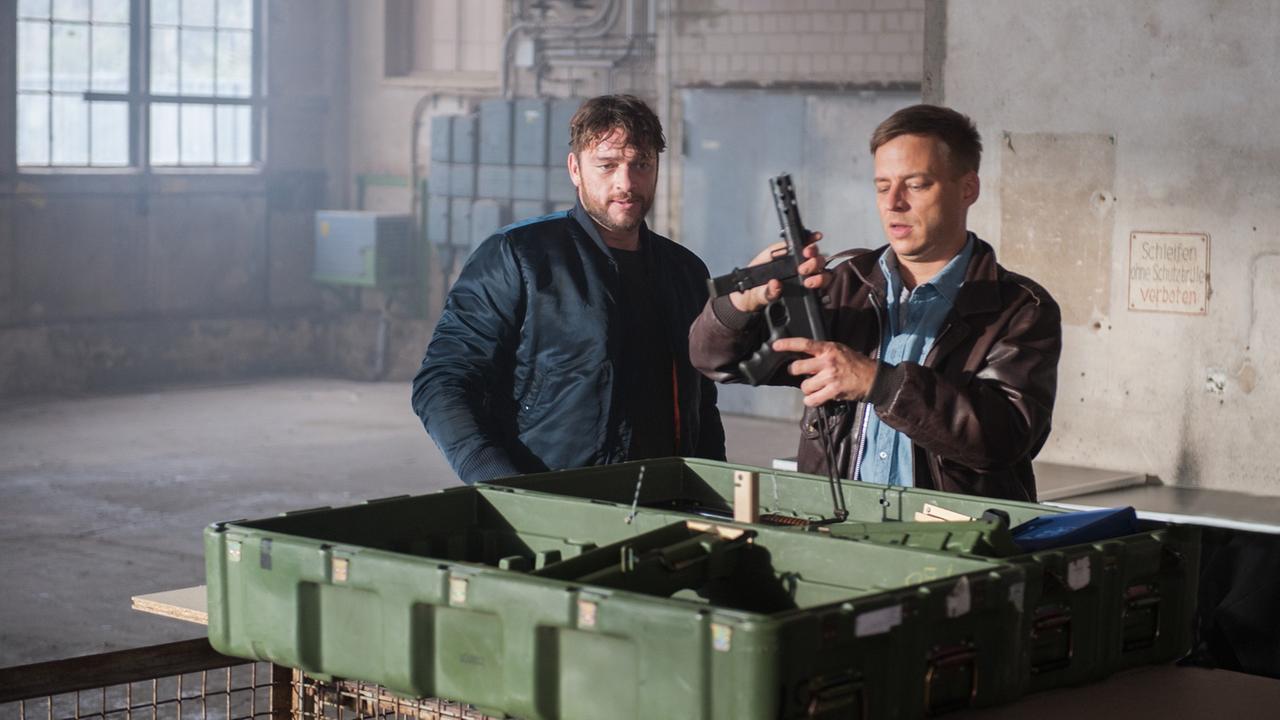 "Dengler - Die schützende Hand": Dengler (Ronald Zehrfeld) und Marius Brauer (Tom Wlaschiha) stehen in einer alten Fabrikhalle vor einem geöffneten Waffenkoffer. Brauer hantiert mit einem Gewehr.