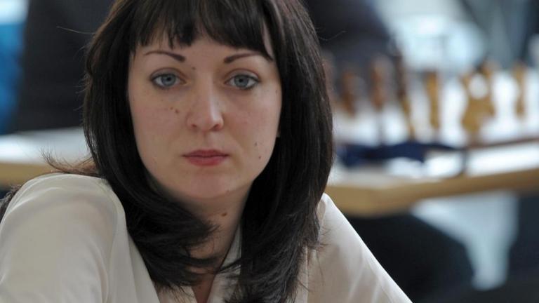 Eine junge Frau sitzt an einem Schachbrett und schaut in diei Kamera. Sie hat dunkle schulterlange Haare. Eine junge Frau sitzt an einem Schachbrett und schaut in diei Kamera. Sie hat dunkle schulterlange Haare.
