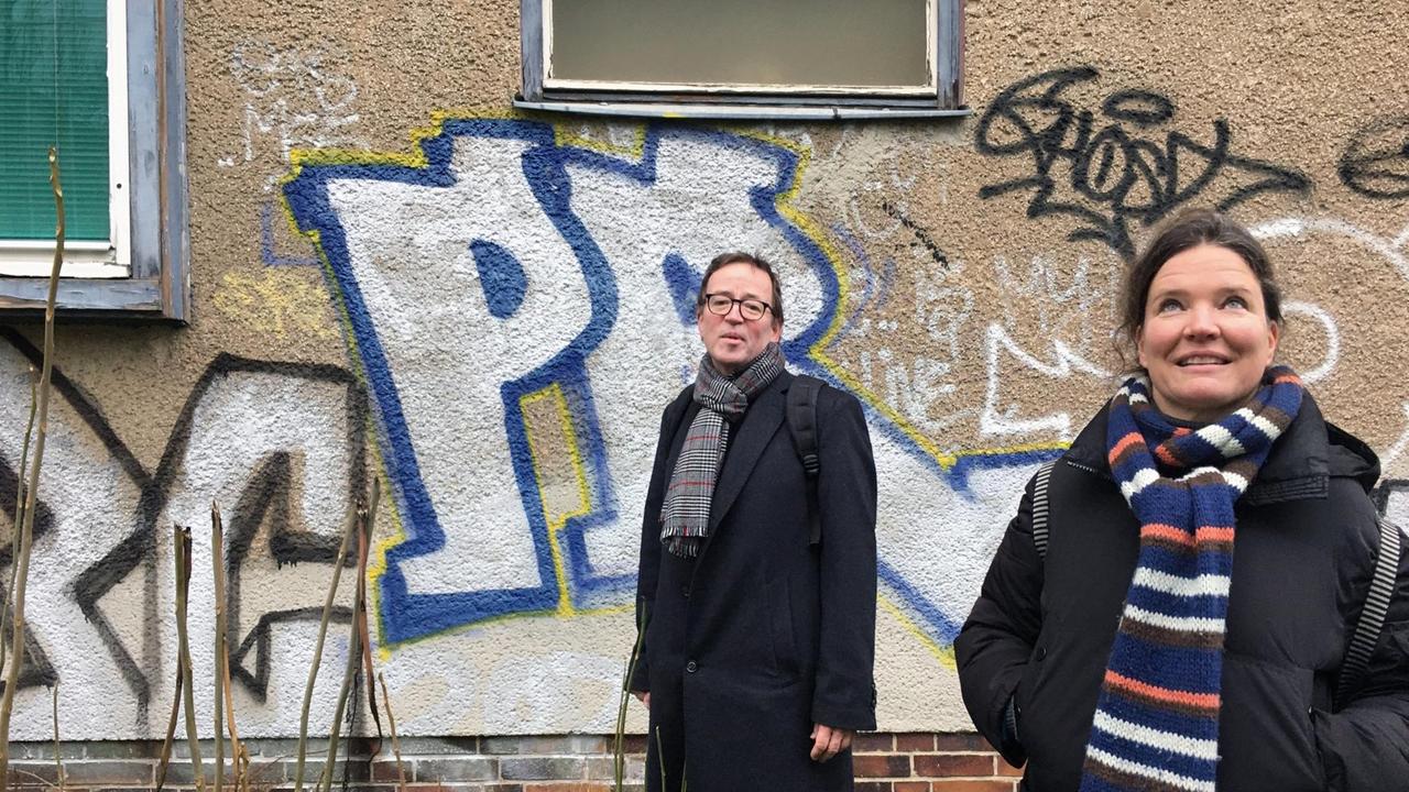 Die Urbanistin Turit Fröbe steht zusammen mit Eckhard Roelcke vor einer, mit Graffiti besprühten, Fasade.