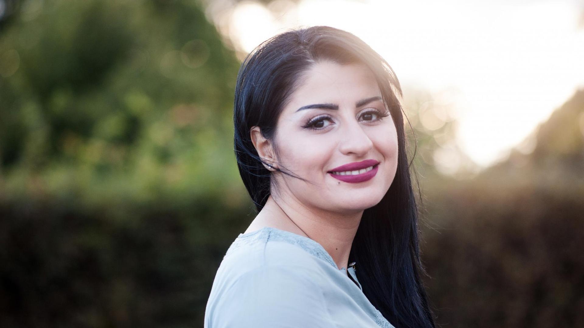 Ein Porträtfoto von Rana Ahmad, eine junge Frau mit langem schwarzem Haar blickt lächelnd über ihre Schulter in die Kamera Ein Porträtfoto von Rana Ahmad, eine junge Frau mit langem schwarzem Haar blickt lächelnd über ihre Schulter in die Kamera