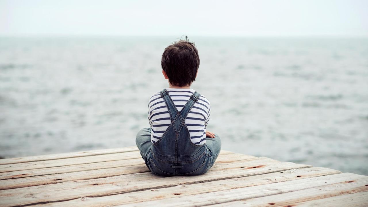 Auf einem Steg sitzt ein kleiner Junge und schaut aufs Meer. Er sieht einsam aus.
