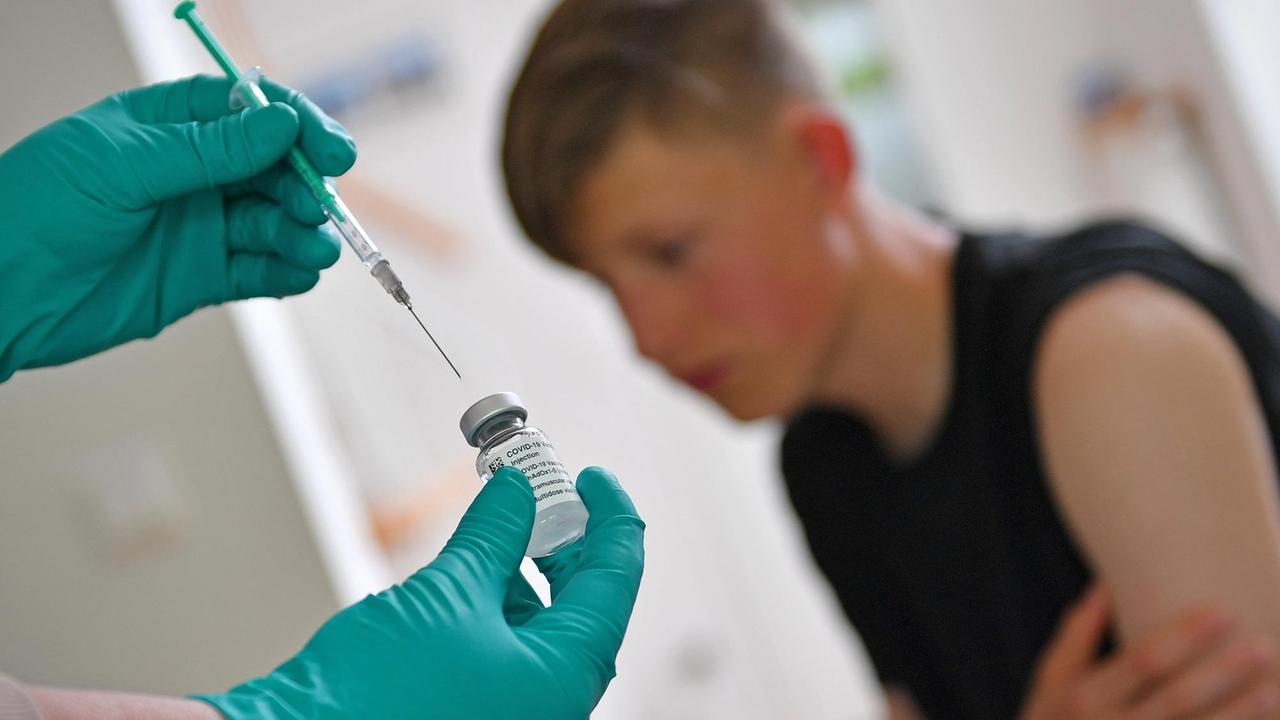 Ein Jugendlicher kurz vor einer Impfung, im Vordergrund sind Hände in Handschuhen zu sehen, die eine Spritze halten. Ein Jugendlicher kurz vor einer Impfung, im Vordergrund sind Hände in Handschuhen zu sehen, die eine Spritze halten.