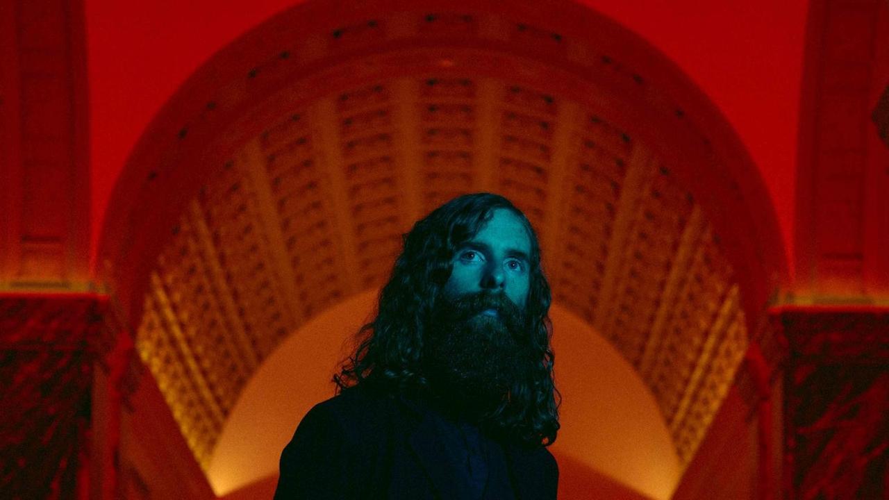 Saintseneca mit Vollbart und langem Haar steht in einen rot ausgeleuchtetem Kirchenschiff. Saintseneca mit Vollbart und langem Haar steht in einen rot ausgeleuchtetem Kirchenschiff.