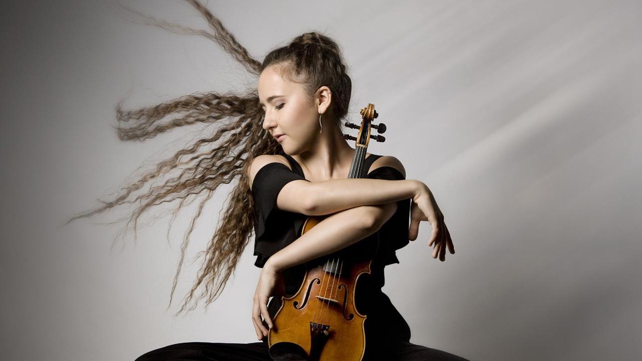 Die Geigerin sitzt mit hochgeschlagenem Bein und Instrument auf einem Drehstuhl, die langen Haare wirbeln durch die Luft. Die Geigerin sitzt mit hochgeschlagenem Bein und Instrument auf einem Drehstuhl, die langen Haare wirbeln durch die Luft.