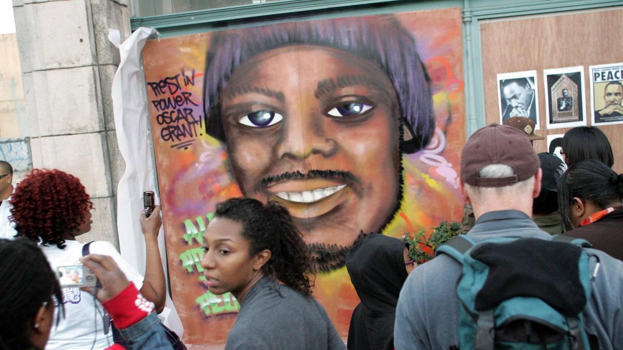 Schwarze und weiße Amerikaner betrachten ein Wandgemälde an einer U-Bahnstation, das einen jungen Afroamerikaner zeigt.