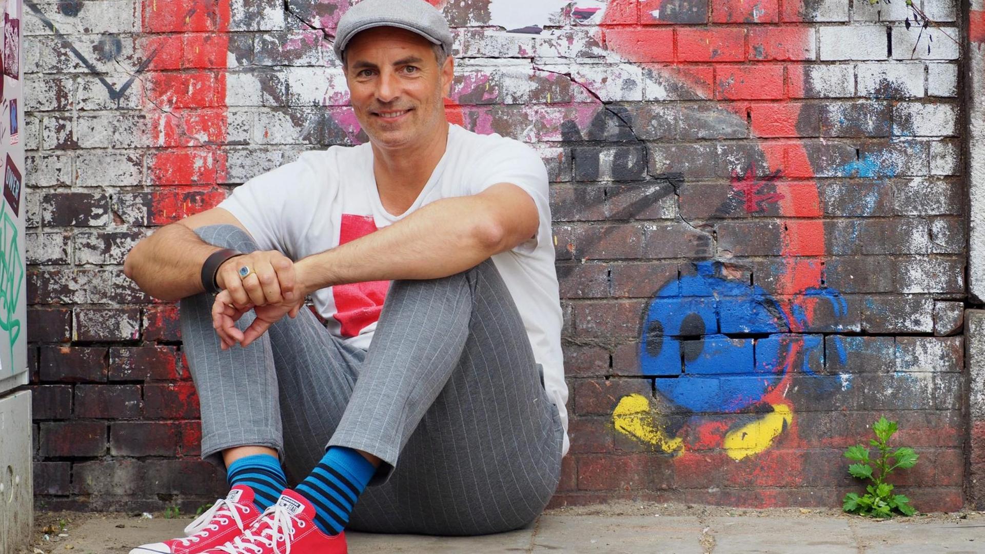Nils Mohl sitzt vor einer mit Graffito besprühten Backsteinmauer. Nils Mohl sitzt vor einer mit Graffito besprühten Backsteinmauer.