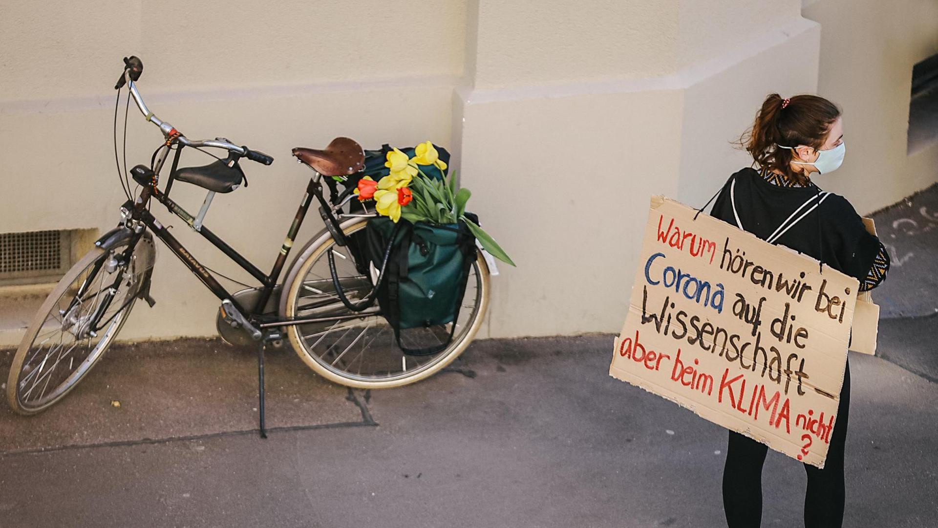 Eine junge Frau mit Mundschutz trägt ein Papschild auf dem Rücken auf dem steht: "WArum hören wir bei Corona auf die Wissenschaft, aber beim Klima nicht?" Im Hintergrund steht ein Fahrrad mit Blumen. Eine junge Frau mit Mundschutz trägt ein Papschild auf dem Rücken auf dem steht: "WArum hören wir bei Corona auf die Wissenschaft, aber beim Klima nicht?" Im Hintergrund steht ein Fahrrad mit Blumen.
