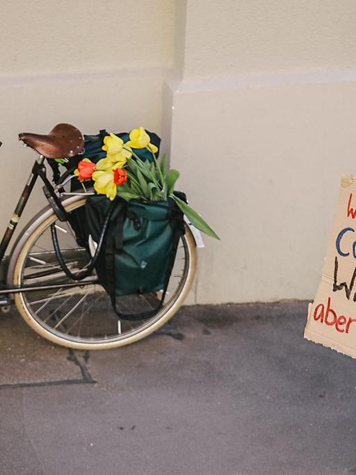 Eine junge Frau mit Mundschutz trägt ein Papschild auf dem Rücken auf dem steht: "WArum hören wir bei Corona auf die Wissenschaft, aber beim Klima nicht?" Im Hintergrund steht ein Fahrrad mit Blumen.