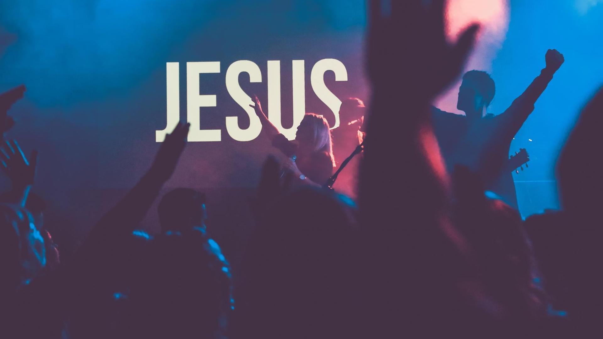 Ein Pop-Konzert in einer Kirche, mit blauem Licht und tanzenden Silhouetten. Im Hintergrund strahlt ein weißer "Jesus"-Schriftzug. Ein Pop-Konzert in einer Kirche, mit blauem Licht und tanzenden Silhouetten. Im Hintergrund strahlt ein weißer "Jesus"-Schriftzug.