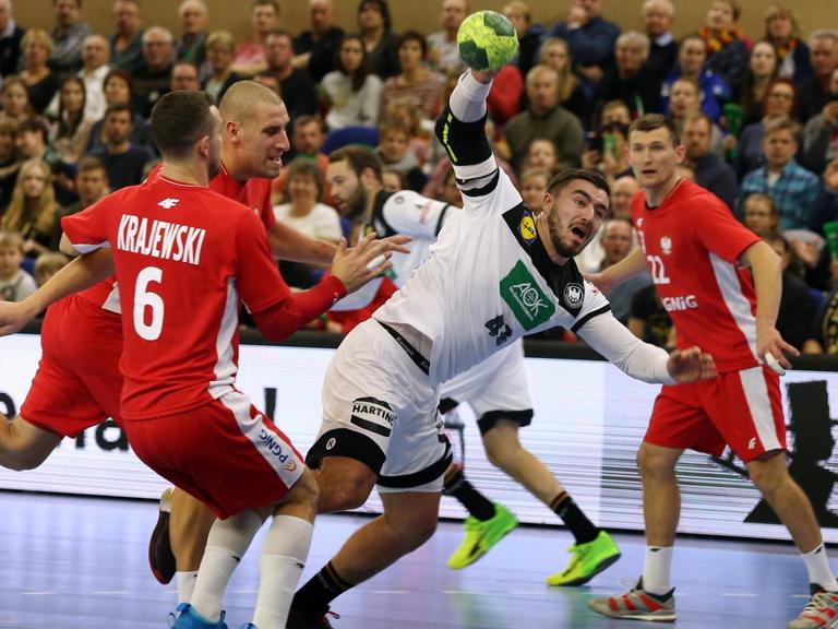 Der deutsche Handball-Spieler Jannik Kohlbacher ist in der Bildmitte zu sehen, wie er sich gegen mehrere Spieler der polnischen Nationalmannschaft durchsetzt.