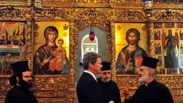 Bundesaußenminister Guido Westerwelle steht 2011 begleitet von drei orthodoxen Priestern im Dreifaltigkeitskloster, das zum Priesterseminar von Chalki gehört. An den Wänden sind zahlreiche goldverzierte Ikonen und Heiligenbilder zu sehen.