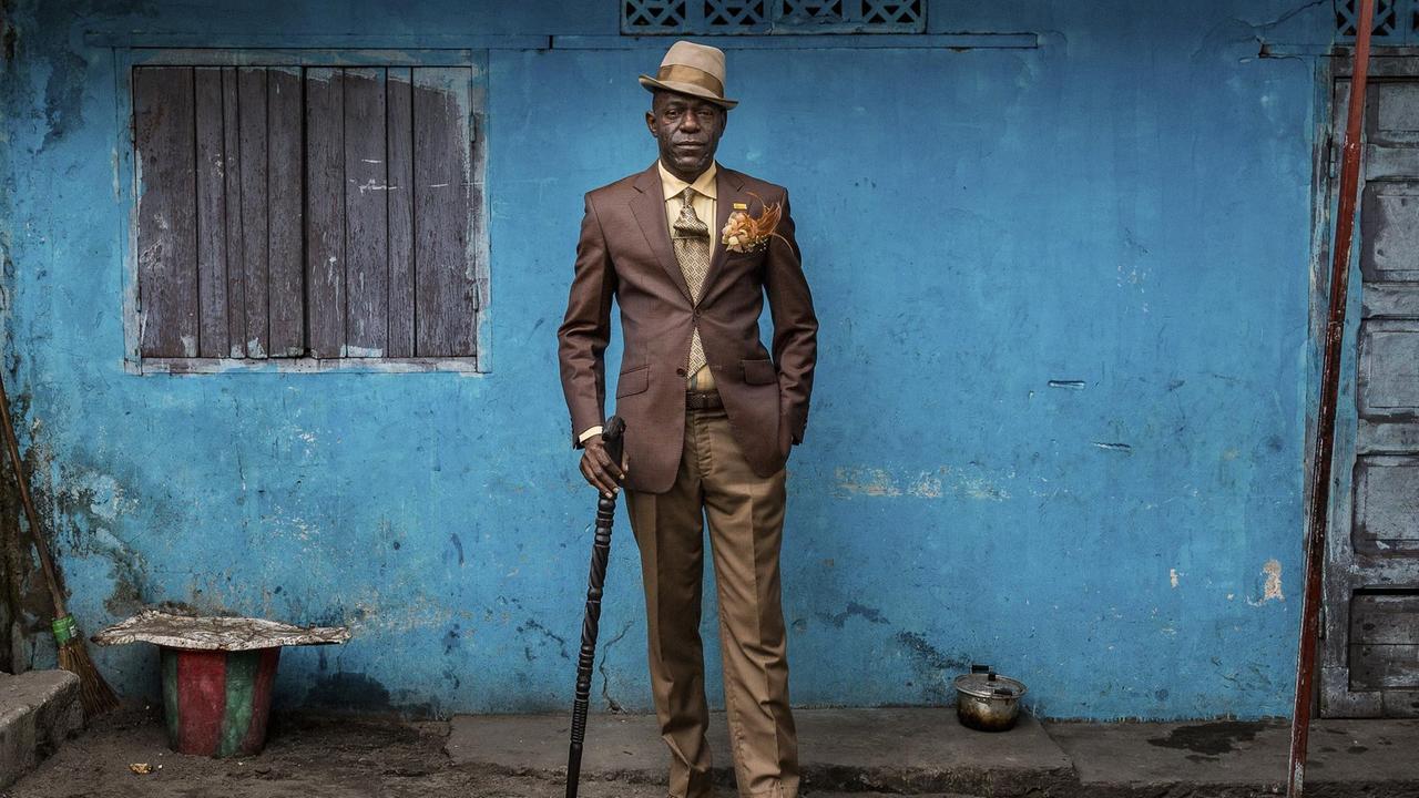 Ein kongolesischer Sapeur steht in elegantem Anzug vor einer blauen Wand.